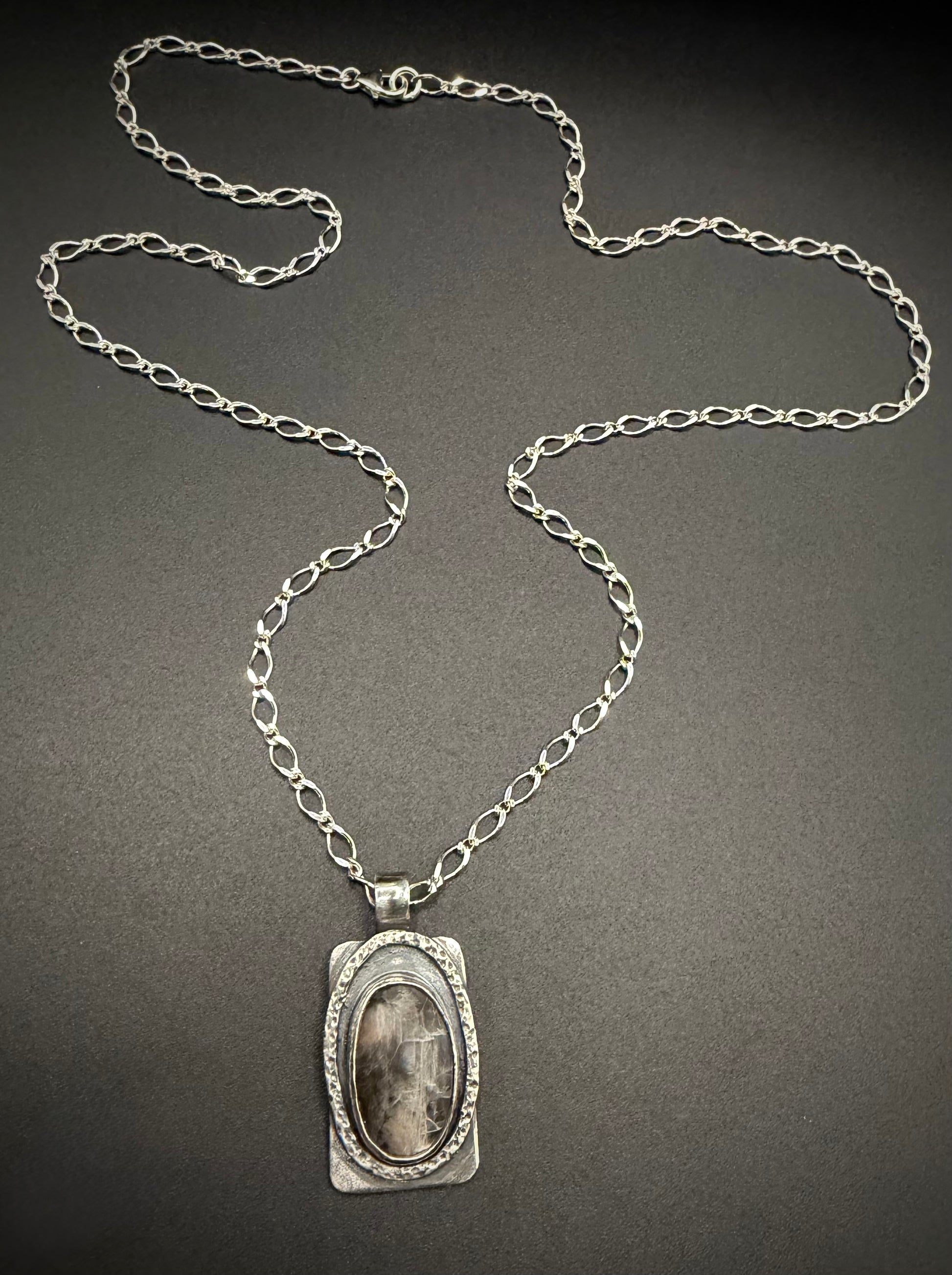 A handcrafted rectangular sterling silver pendant on a long link chain, resting against a dark background. The pendant features a central, vertically oriented oval black moonstone cabochon enclosed by a deeply textured, layered silver bezel.
