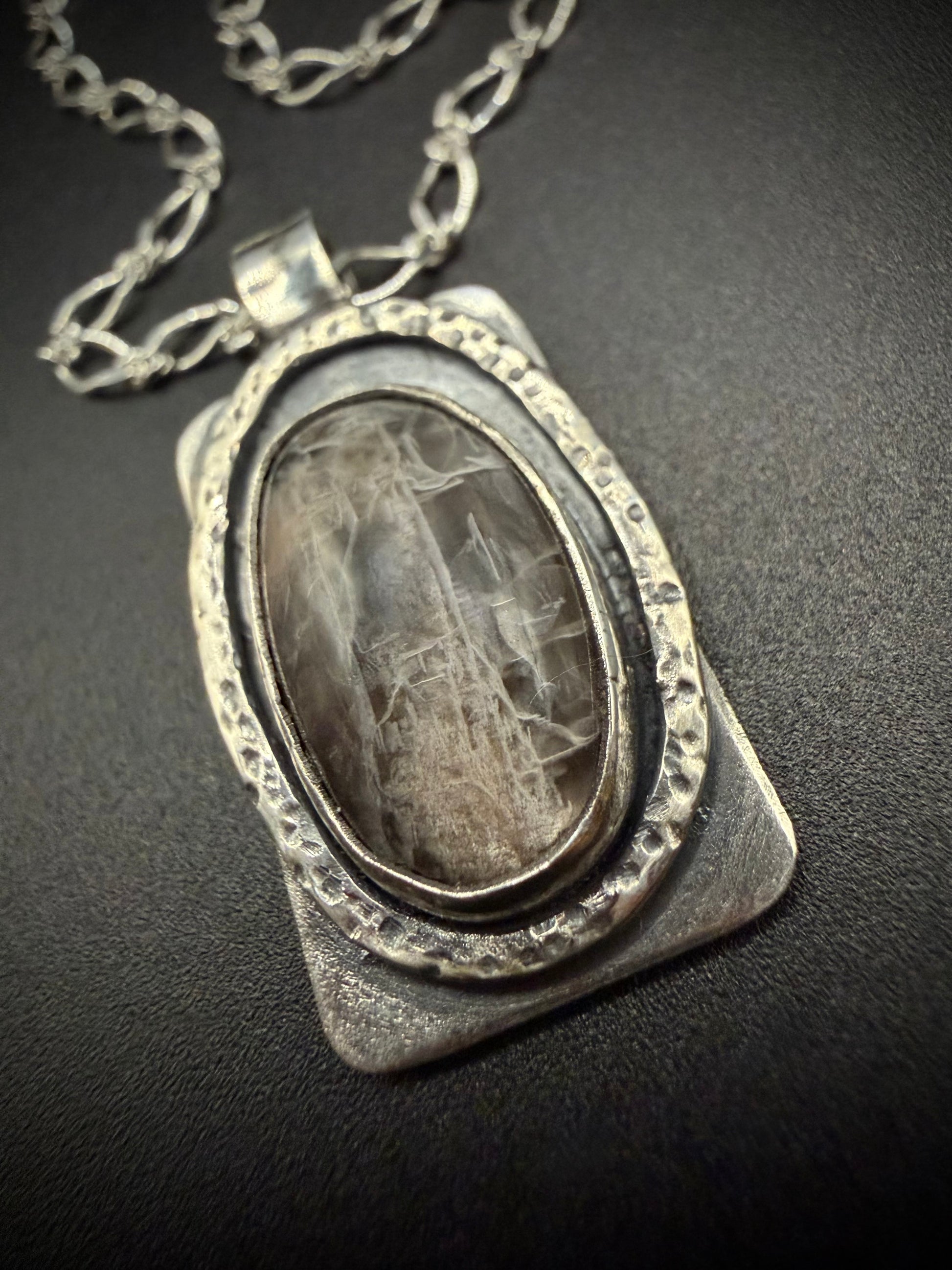 A close-up shot of a handcrafted rectangular sterling silver pendant resting on a dark surface, featuring a large oval black moonstone with visible internal fracturing, set within a layered, deeply hammered oval bezel on a textured silver backplate.