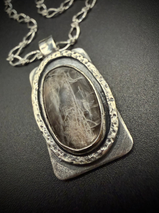 A close-up shot of a handcrafted rectangular sterling silver pendant resting on a dark surface, featuring a large oval black moonstone with visible internal fracturing, set within a layered, deeply hammered oval bezel on a textured silver backplate.