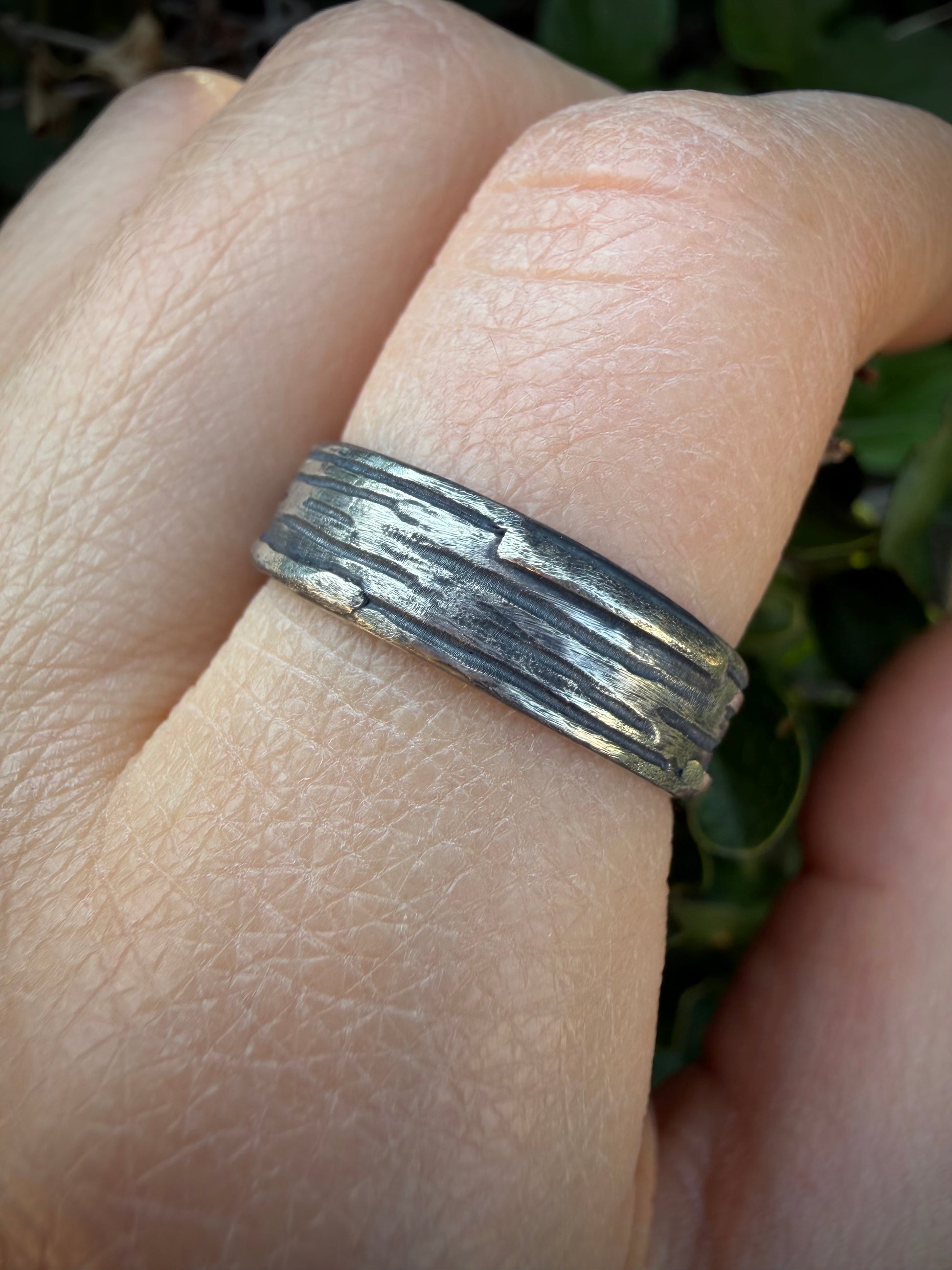 A close-up of a textured silver band ring worn on a person's finger, with a blurred green outdoor background.