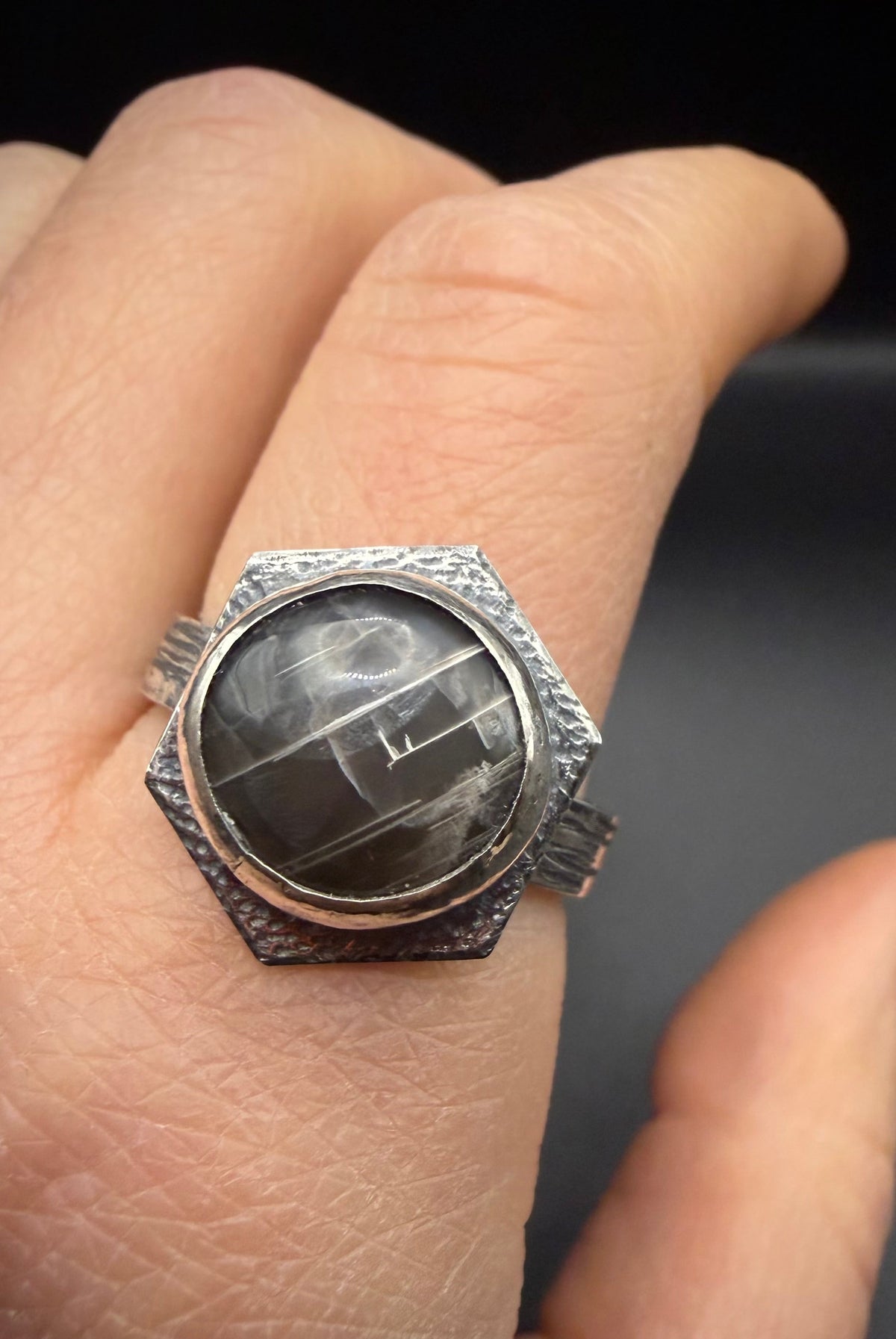 A close-up of a hand wearing a textured silver hexagonal ring with a dark, cloudy stone cabochon.