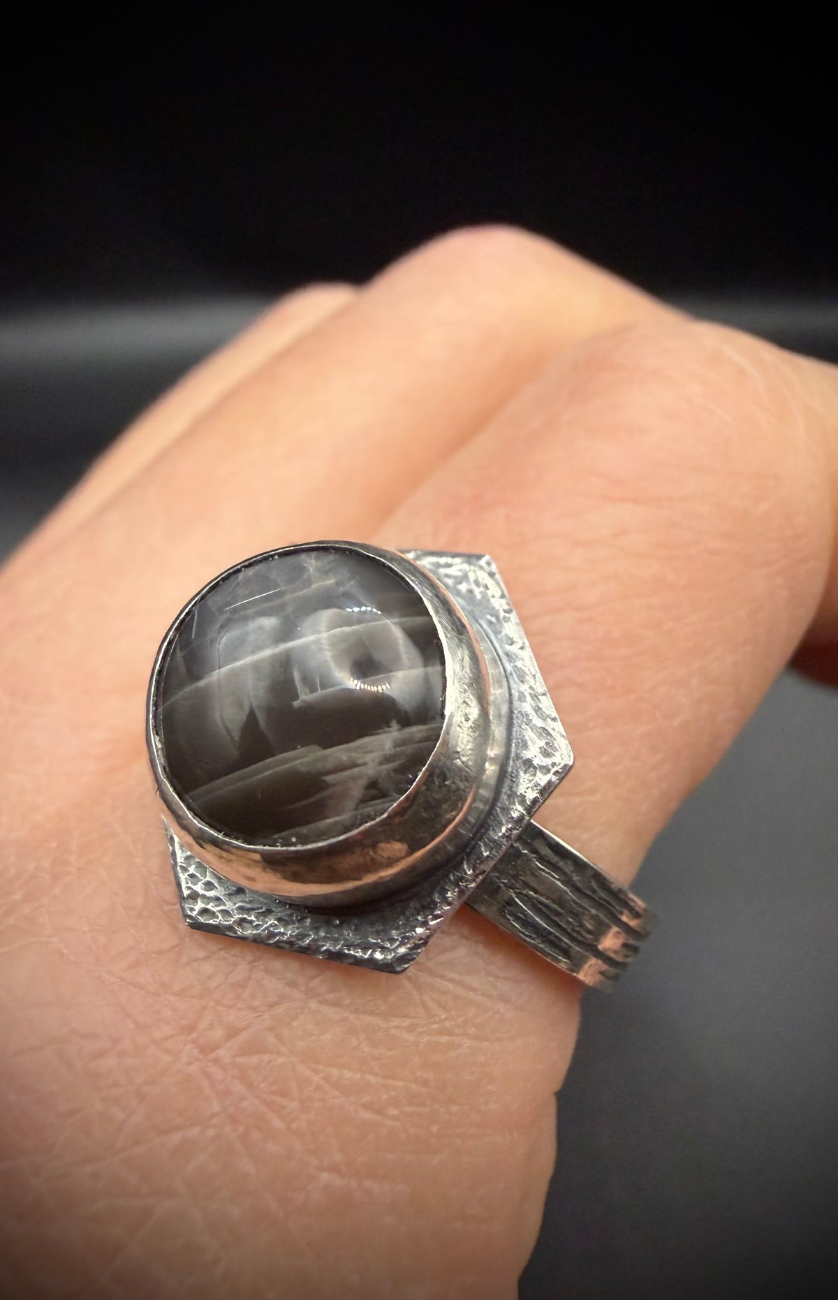 A silver ring with a large round black moonstone, worn on a person's finger against a dark background.