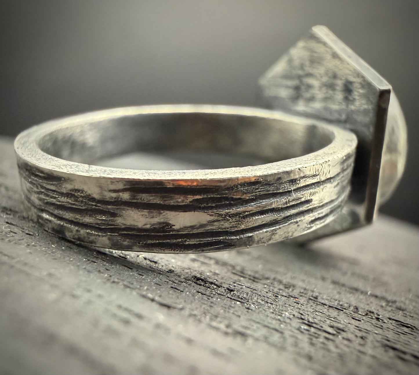 A close-up of a textured silver ring with a geometric detail, resting on a weathered gray wooden surface.