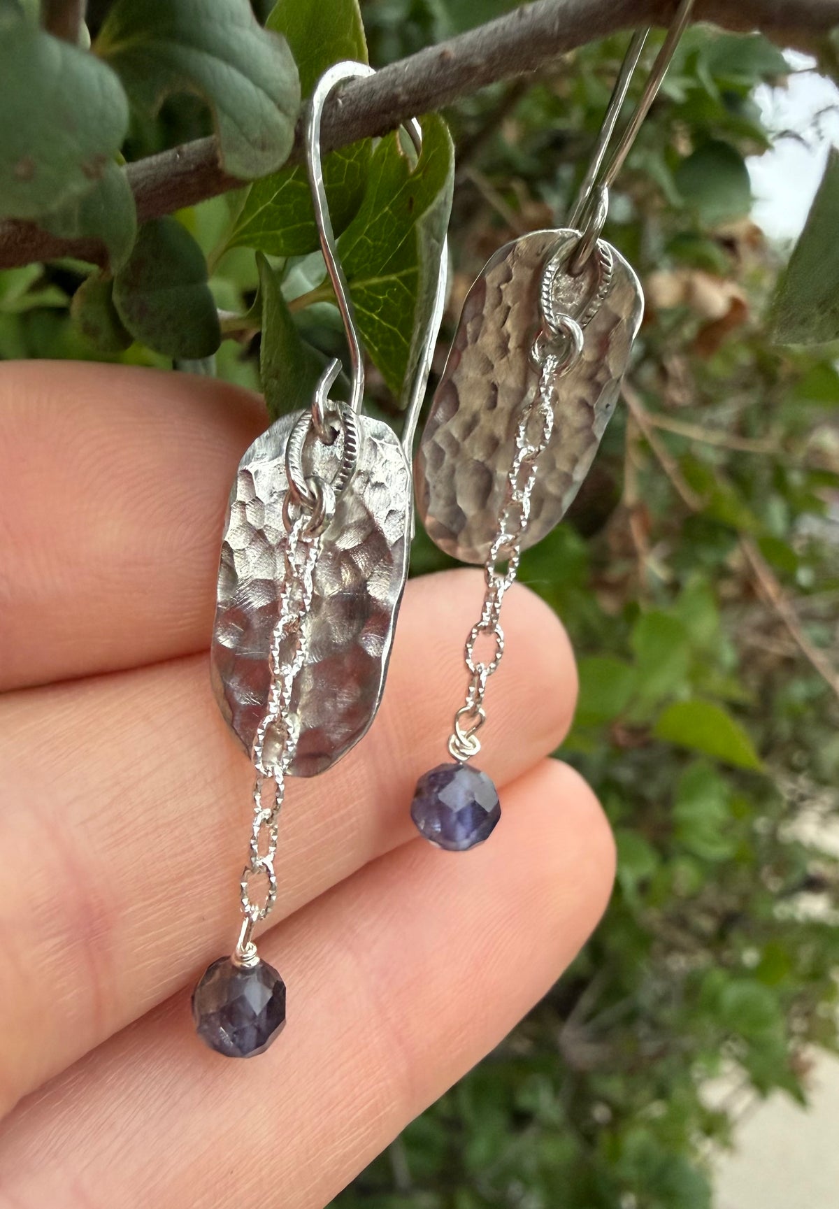 Handmade hammered silver dangle earrings with faceted purple beads, held in a person's hand against a blurred green outdoor background.