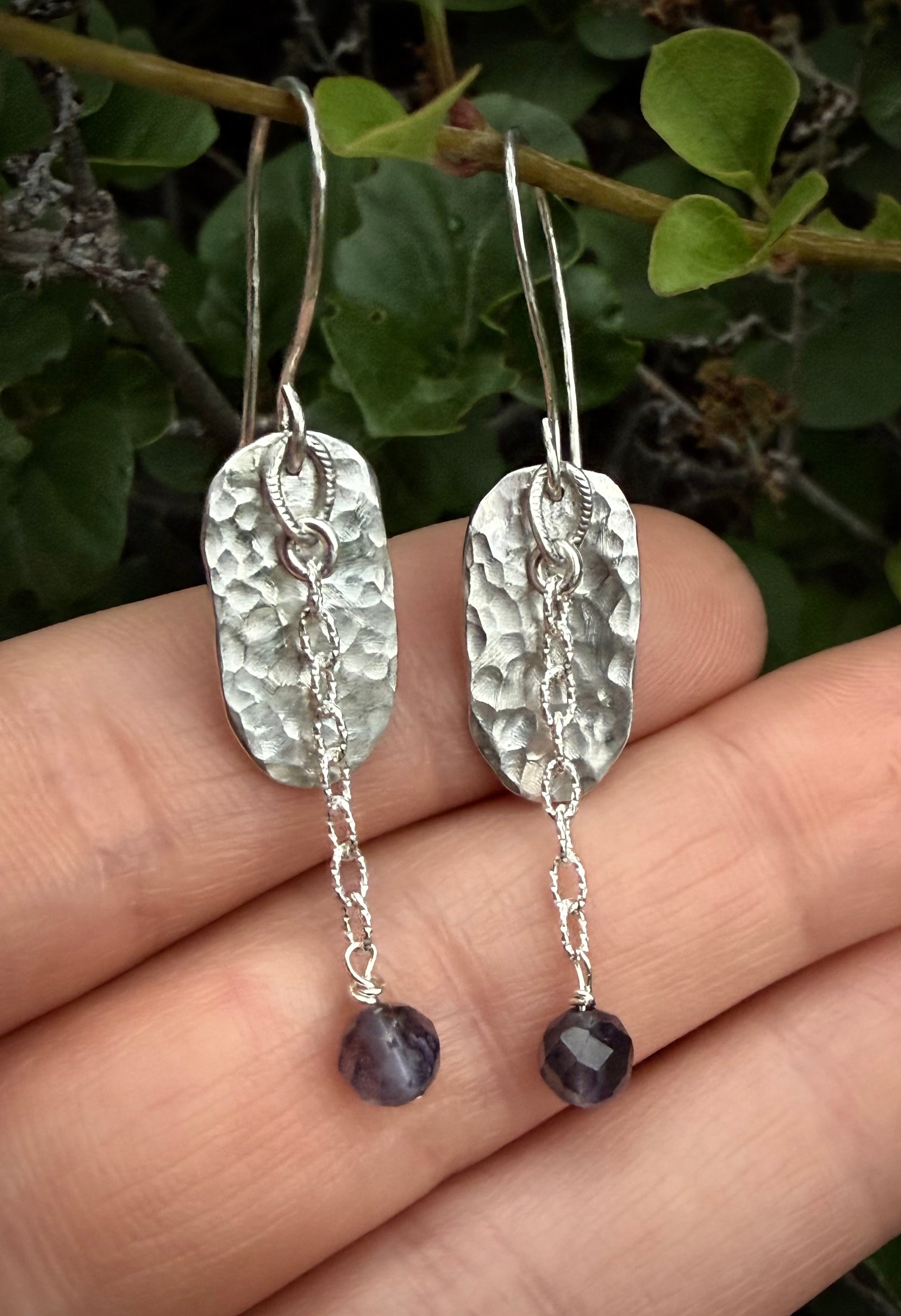 A pair of hammered silver drop earrings with small silver chain links and faceted dark purple beads, shown hanging over an open hand with green leaves in the background.