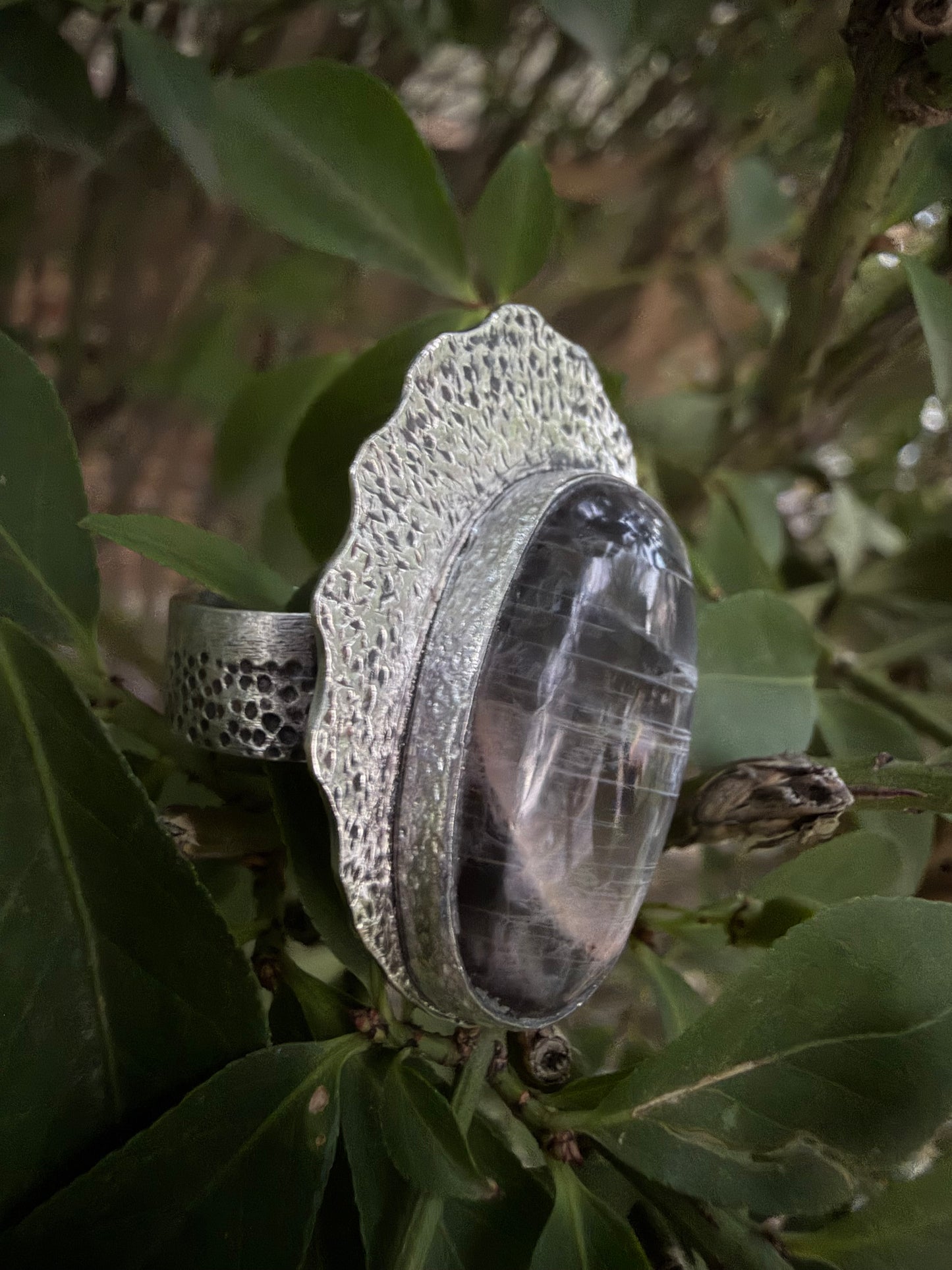 A large silver ring with a scalloped textured setting and a prominent oval dark gray stone, photographed resting on green leaves outdoors.
