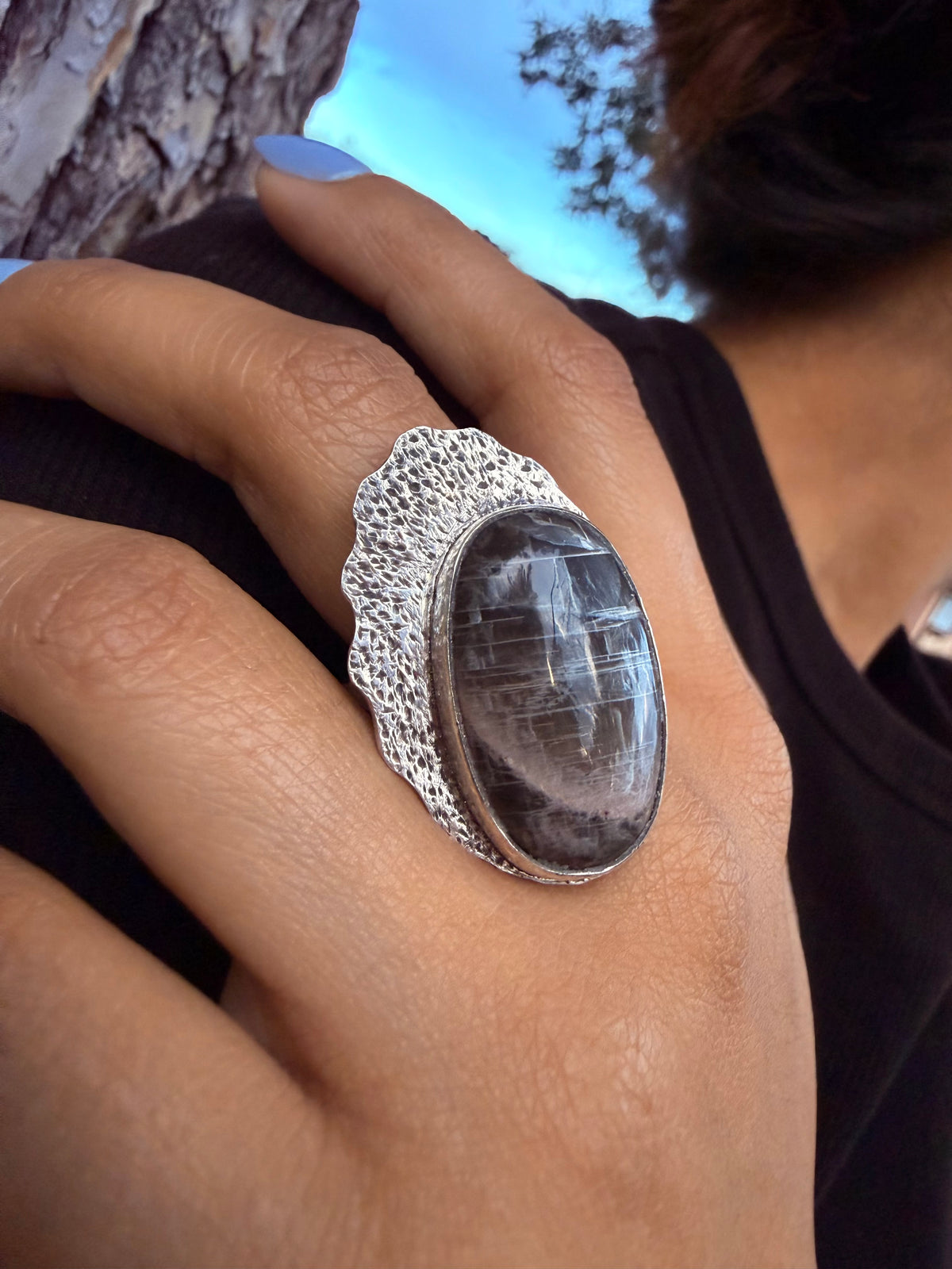A close-up of a hand wearing a large silver ring with an oval dark smoky stone, set against an outdoor background with a tree trunk and blue sky.