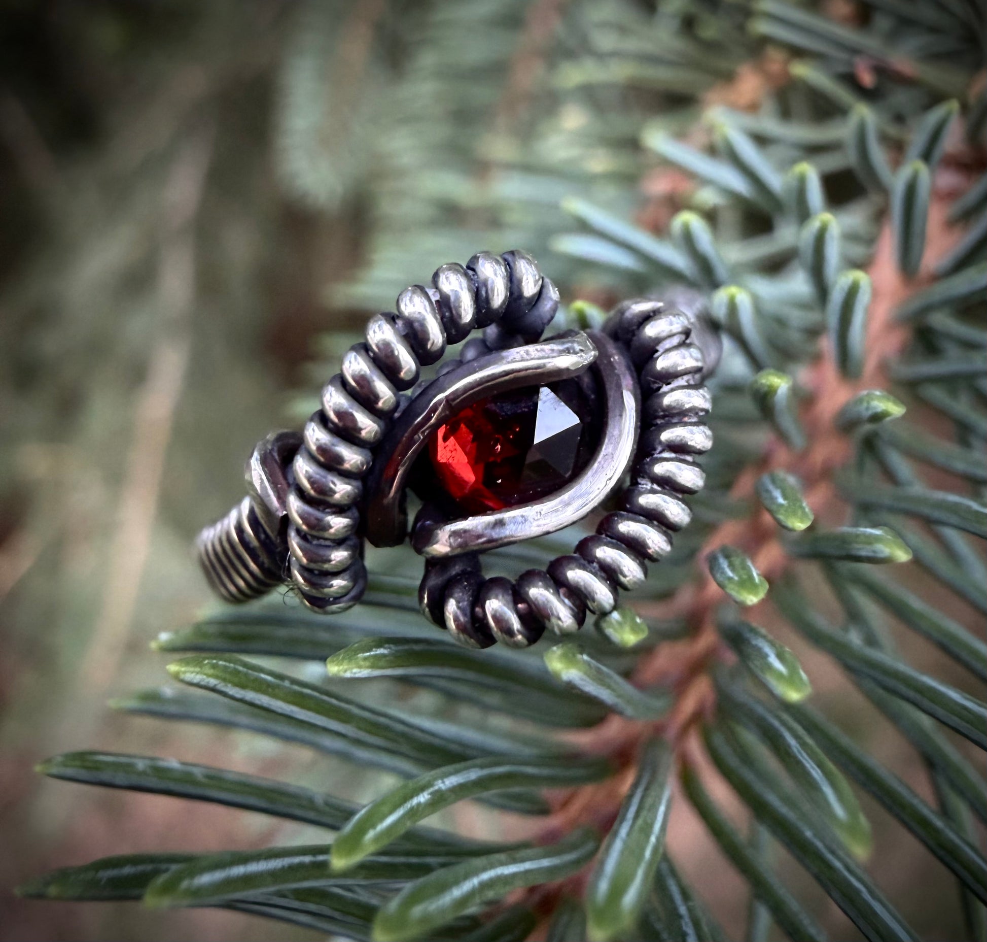 A handmade silver ring with a faceted red gemstone, wrapped in a textured spiral design, resting on green pine needles.