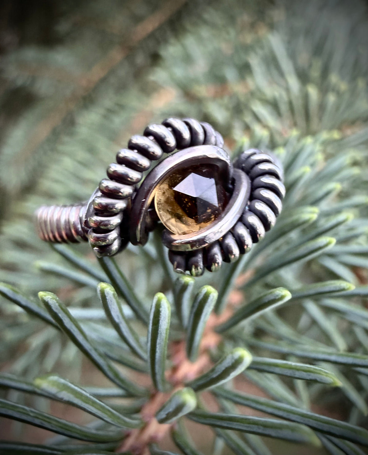 A close-up of a handmade silver ring with a faceted amber gemstone, resting on the needles of a pine tree.