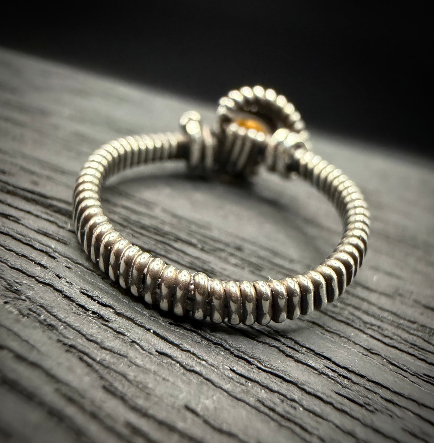 A handmade sterling silver ring with a textured, ribbed band and a small amber-colored stone, resting on a dark wood surface.