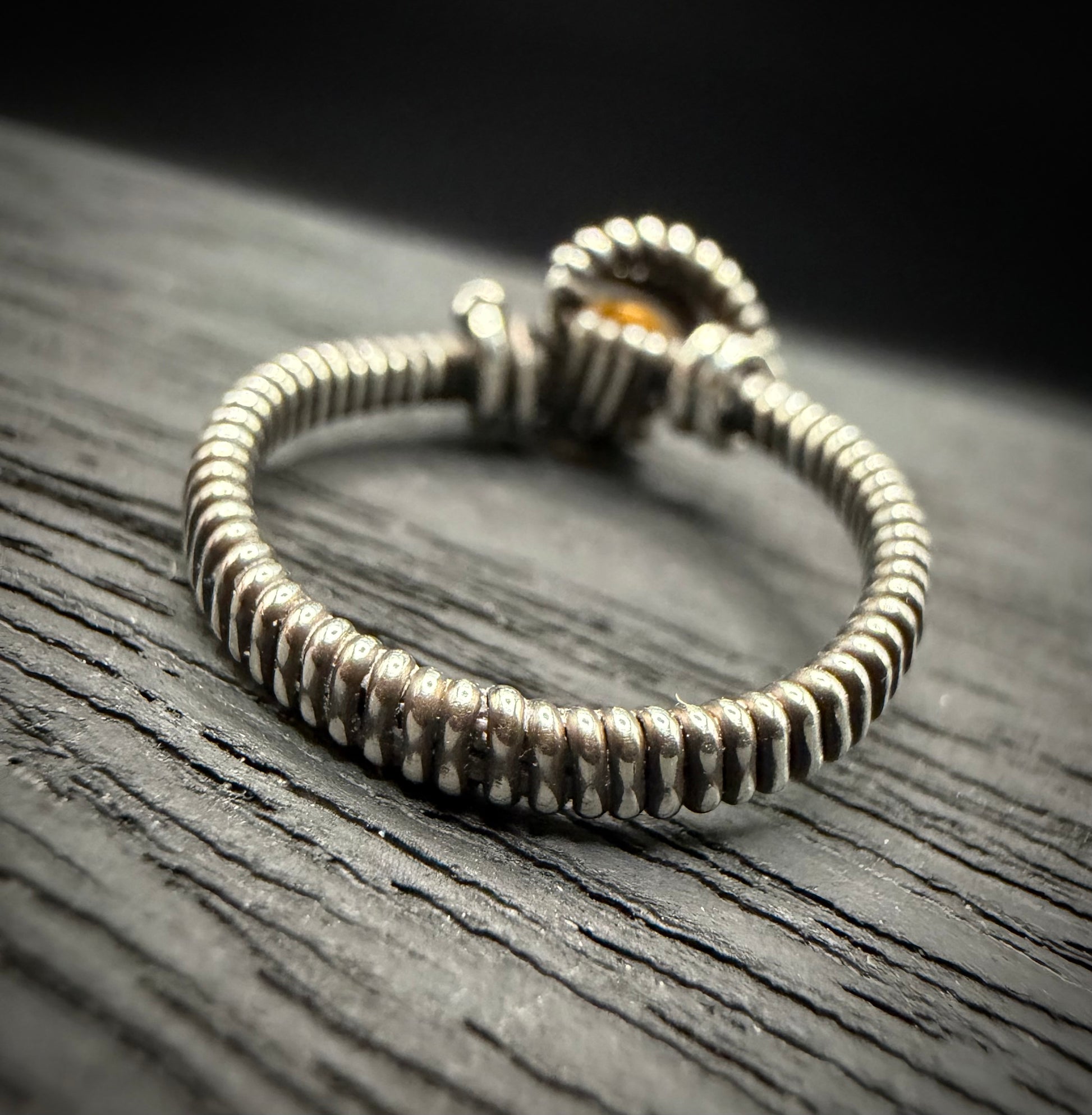 A handmade sterling silver ring with a textured, ribbed band and a small amber-colored stone, resting on a dark wood surface.