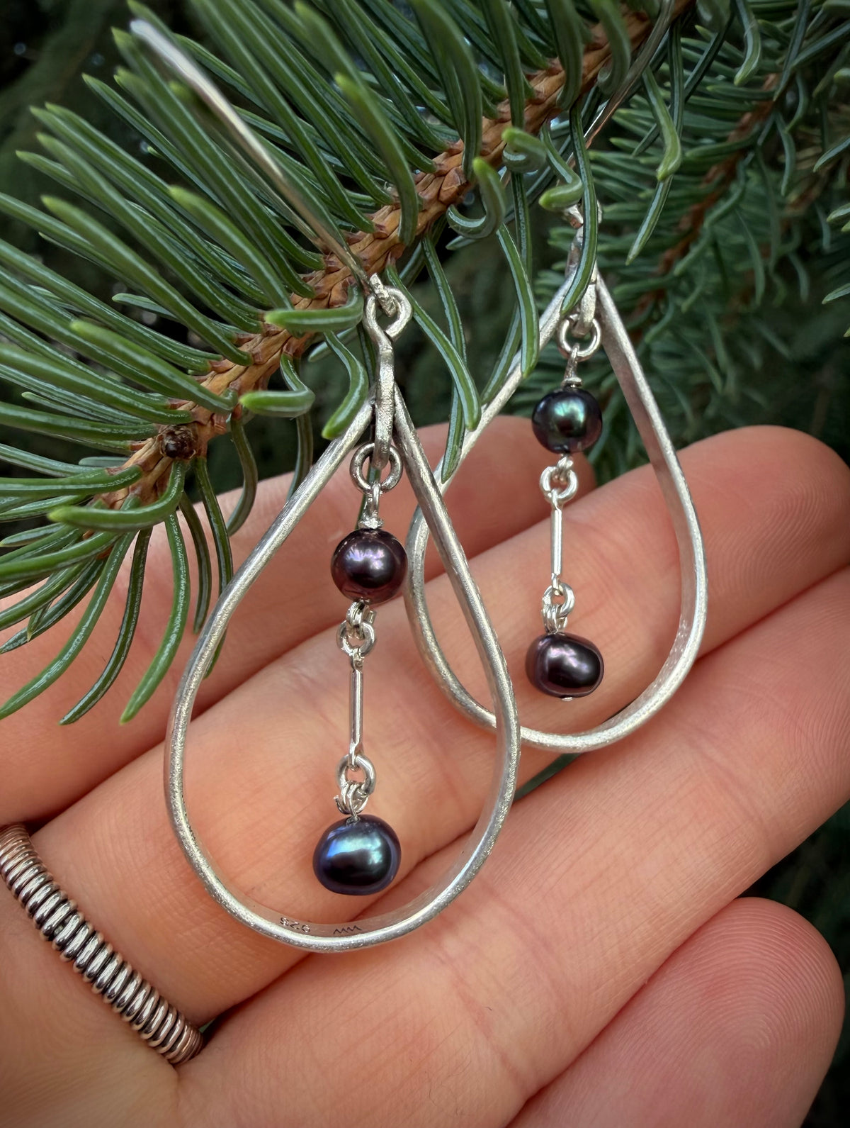 A pair of silver teardrop dangle earrings with small dark pearl beads, held in a person's hand in front of green pine needles.