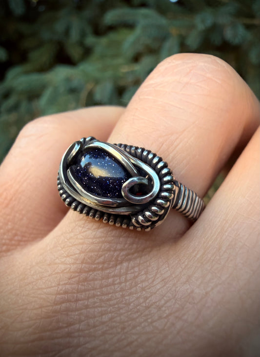 Hand wearing the handcrafted Asteria Ring featuring a glittering blue goldstone set in sterling silver wirework against a blurred green background.