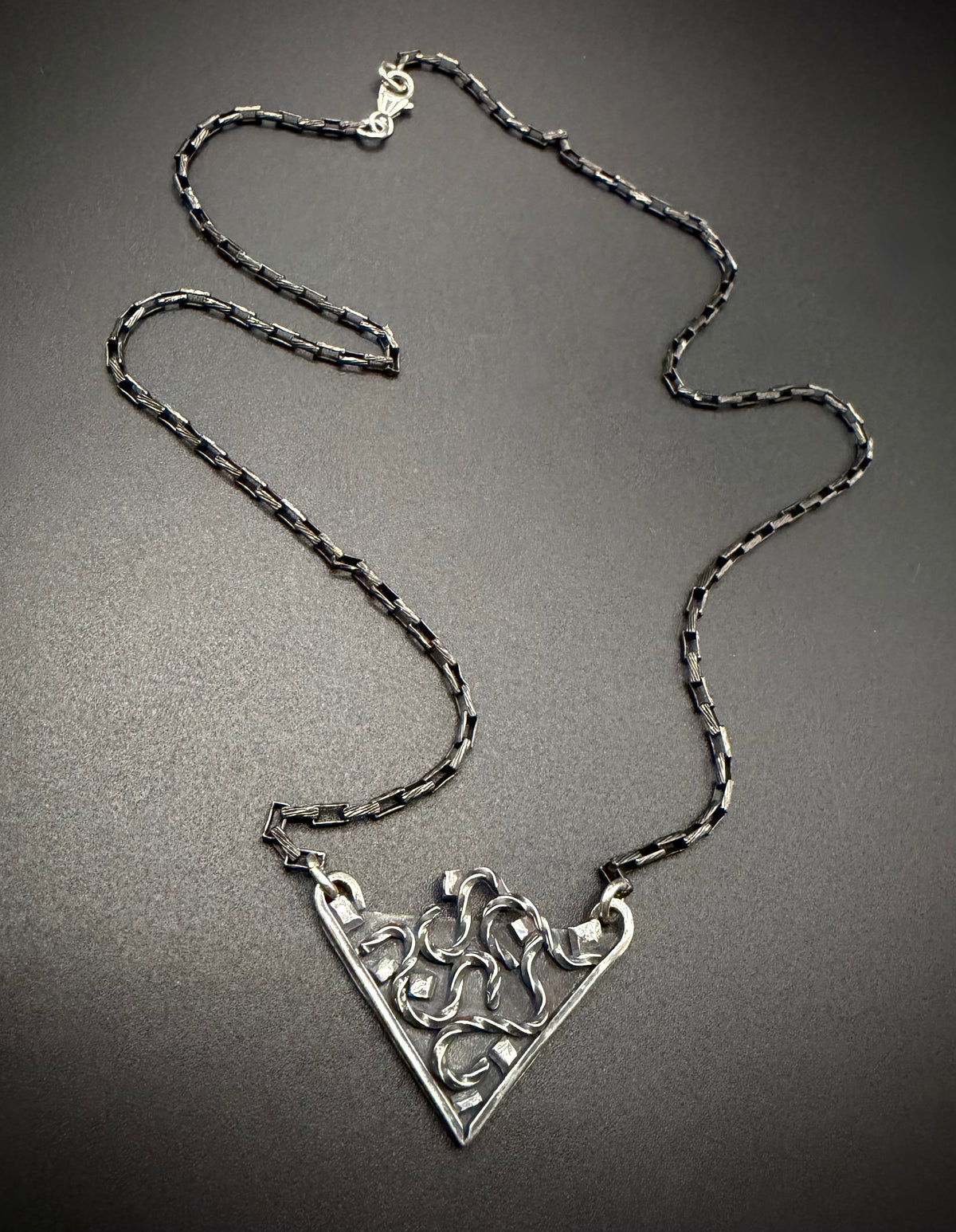 A silver-toned necklace featuring a textured, openwork triangular pendant and a matching chain, displayed on a dark gray background.
