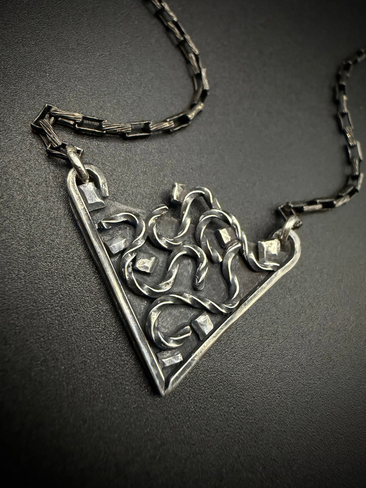 A silver-toned triangular pendant necklace with an intricate openwork knot design, displayed on a textured dark gray background.