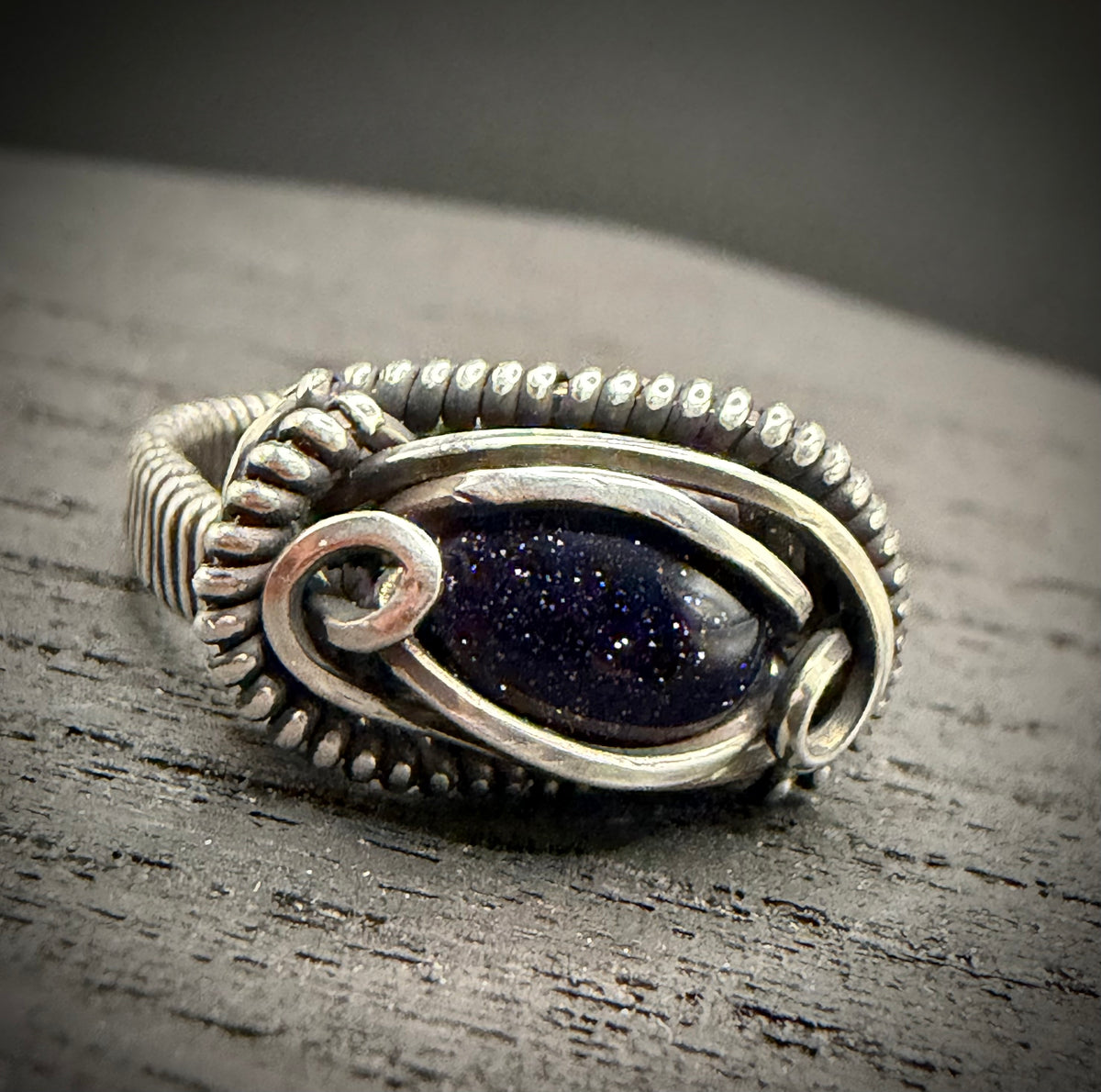 Handcrafted Asteria Ring featuring a shimmering blue goldstone framed by intricate sterling silver wirework, set on a dark rustic wooden background.