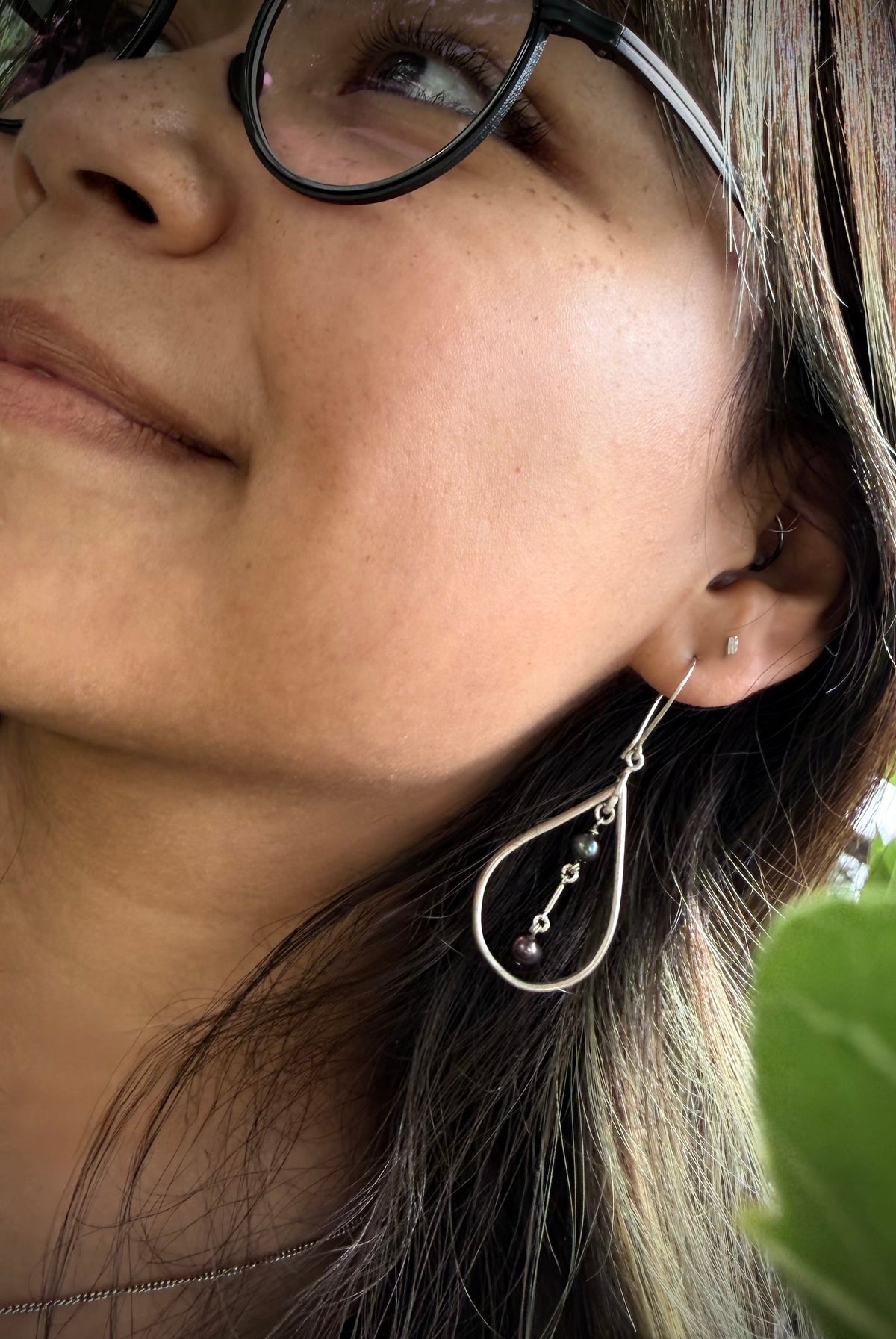 A close-up lifestyle shot of a woman wearing a silver teardrop-shaped dangle earring with small green bead accents. She is wearing black-rimmed glasses and has dark brown hair with blonde highlights, with a green leaf visible in the foreground.