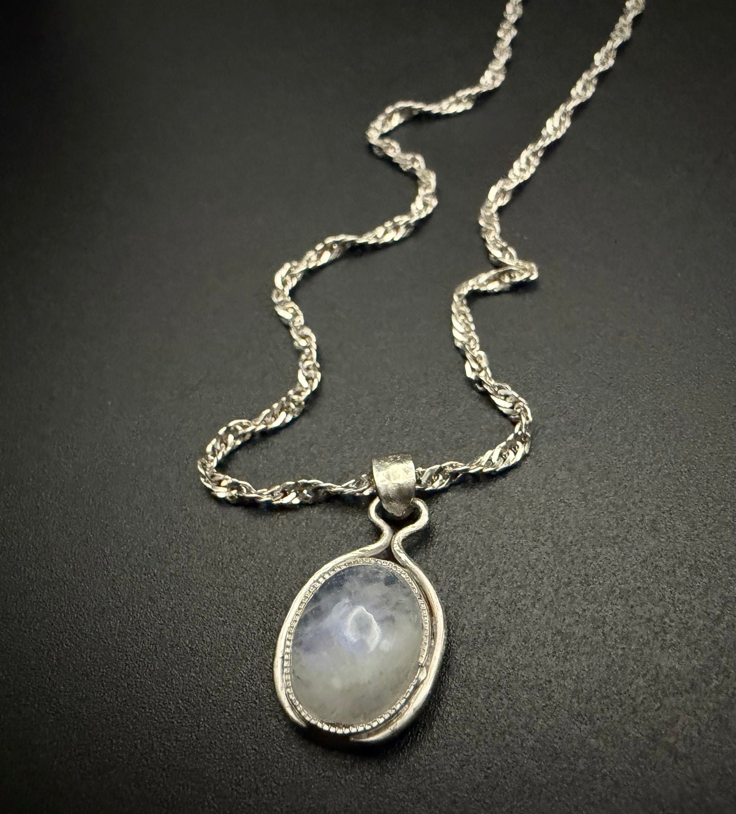 A close-up of a handcrafted sterling silver necklace featuring the Iris Pendant, which holds a luminous, iridescent Moon Stone. The pendant is suspended from a delicate silver rope chain against a dark, textured background.