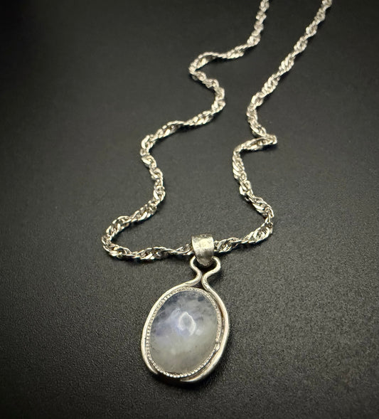 A close-up of a handcrafted sterling silver necklace featuring the Iris Pendant, which holds a luminous, iridescent Moon Stone. The pendant is suspended from a delicate silver rope chain against a dark, textured background.