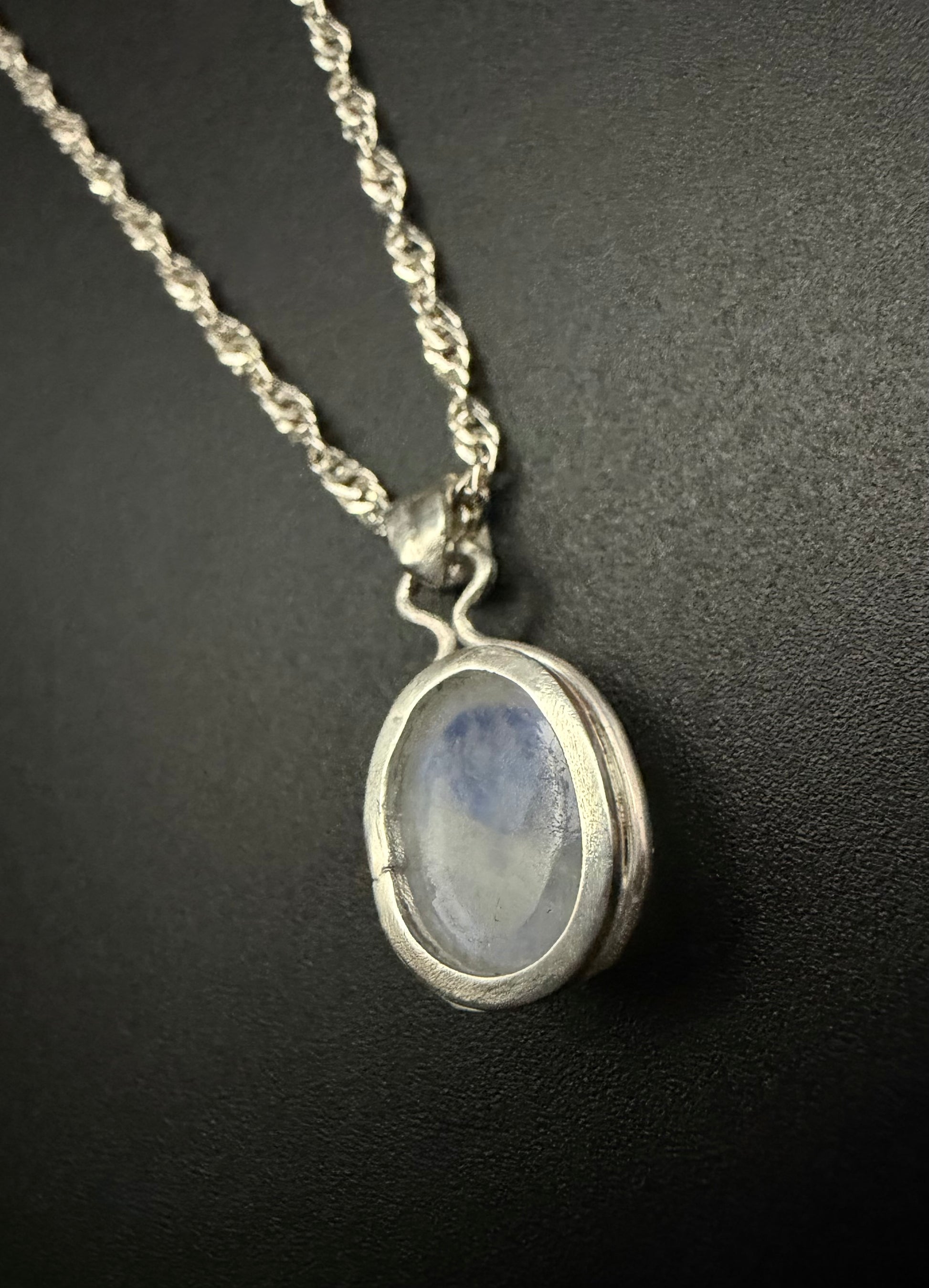 A close-up of a sterling silver necklace featuring the back of the Iris Pendant, which holds a luminous, iridescent Moon Stone. The pendant is suspended from a delicate silver rope chain against a dark, textured background.