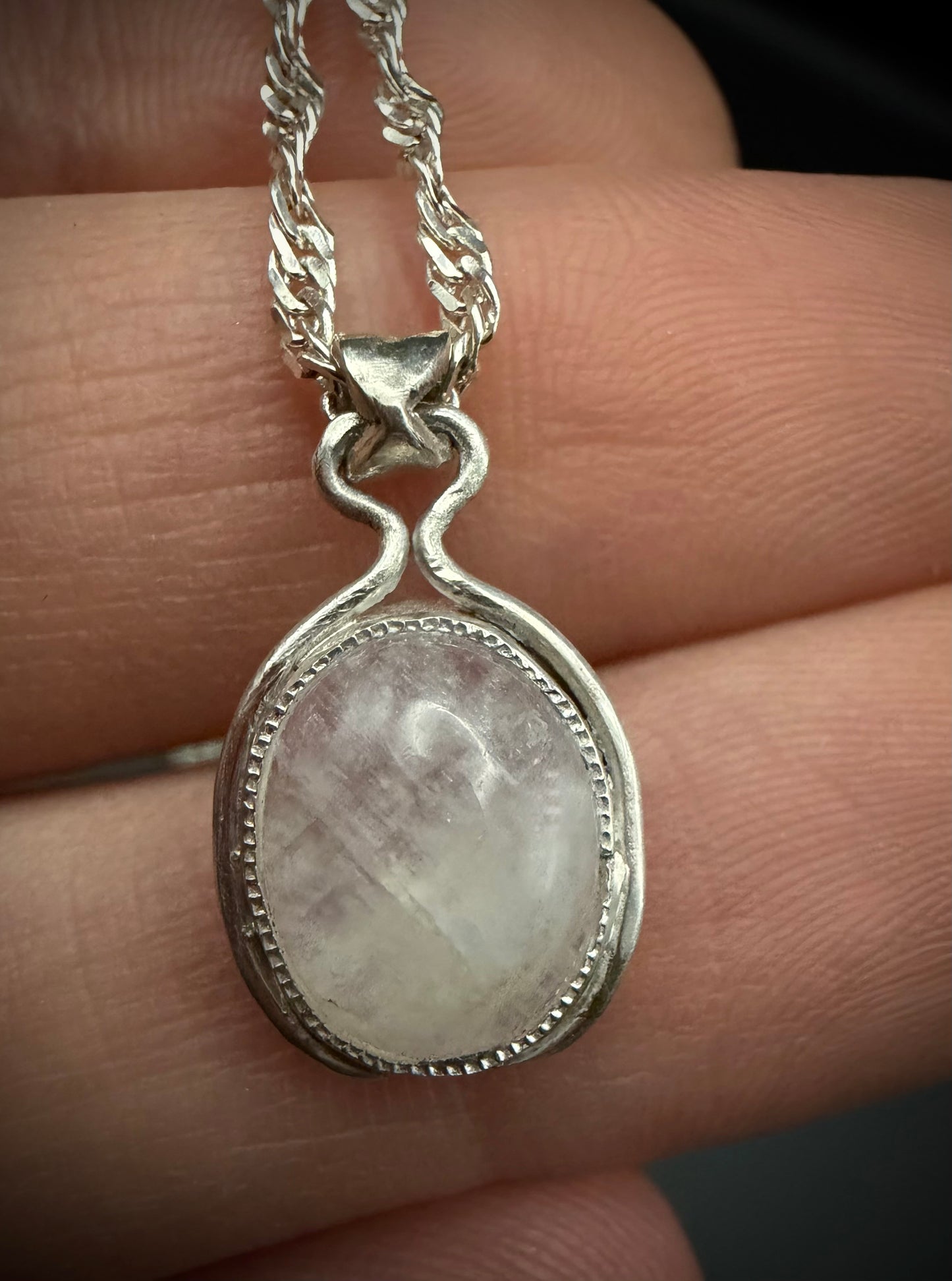 A close-up, hand-held product photograph of the Iris Pendant, featuring a handcrafted sterling silver setting. The oval, iridescent gemstone cabochon is secured with a decorative beaded bezel and suspended from a custom-shaped silver bail on a delicate twisted rope chain.
