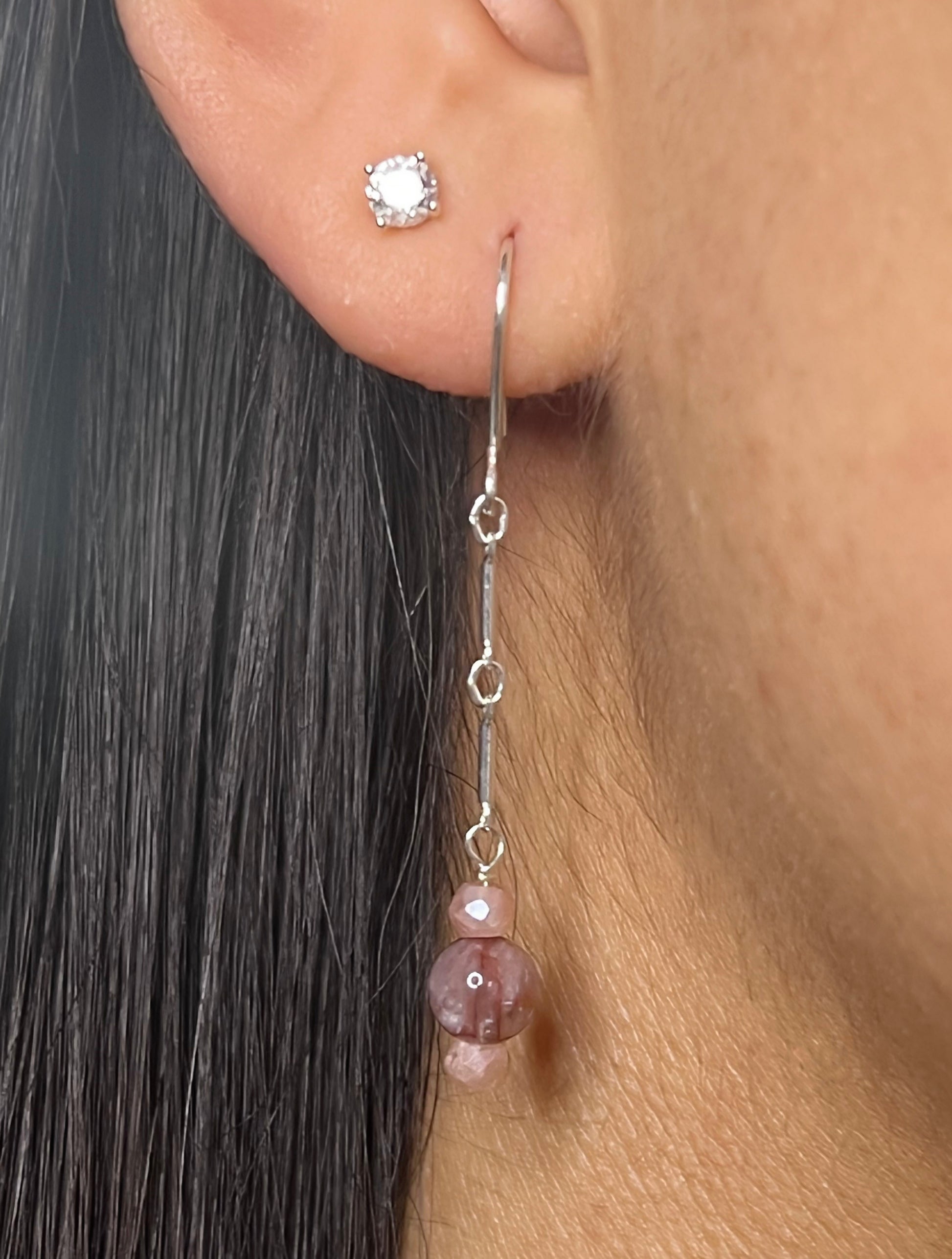 A close-up of a person wearing a handcrafted sterling silver earring featuring a long delicate chain with a stack of faceted peach moonstone and smooth red agate beads.