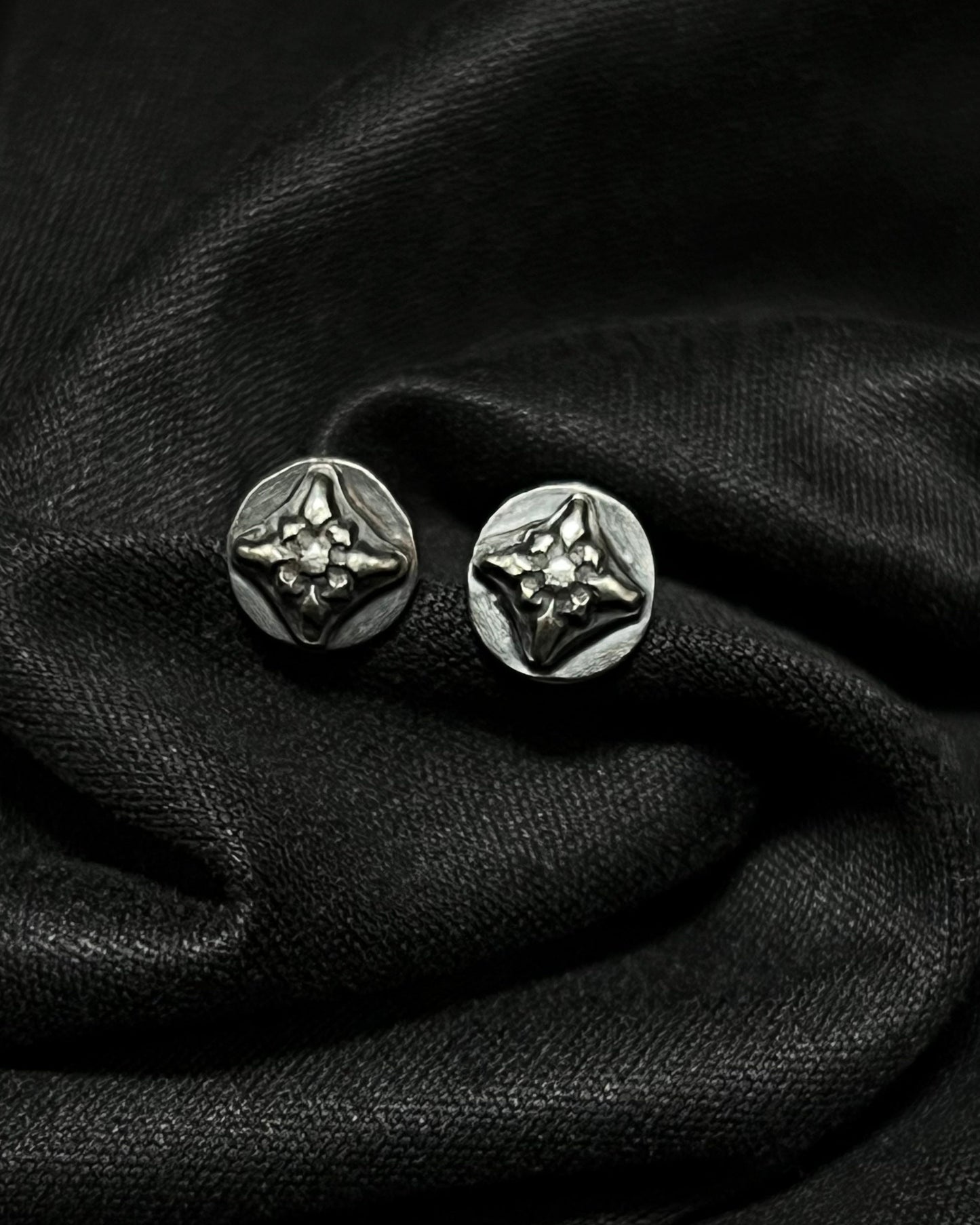 A pair of 7mm Ember Studs from the Ashborn Collection. The sterling silver earrings feature a raised, four-pointed starburst design with a polished finish against a dark, recessed patina, shown on draped black fabric
