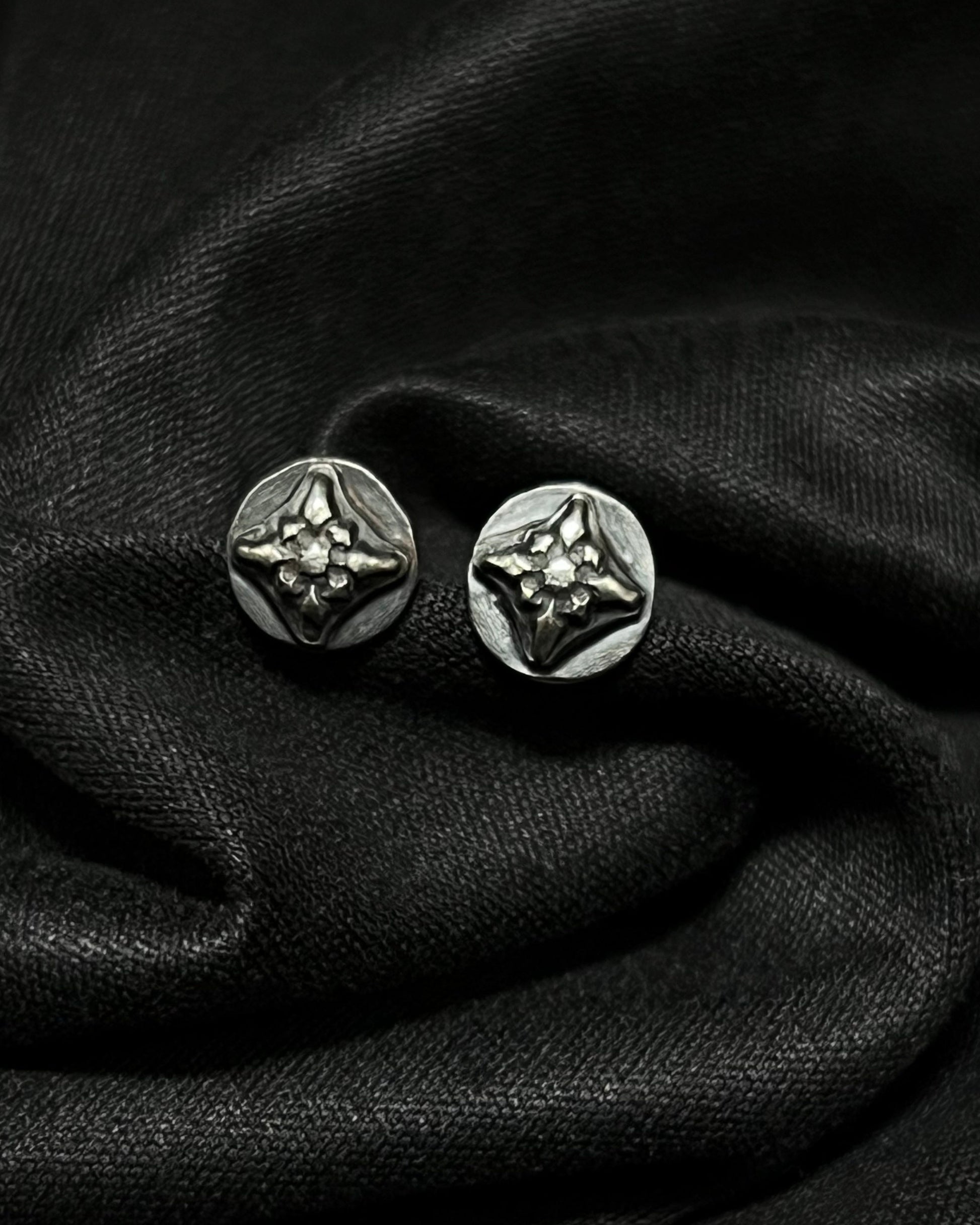 A pair of 7mm Ember Studs from the Ashborn Collection. The sterling silver earrings feature a raised, four-pointed starburst design with a polished finish against a dark, recessed patina, shown on draped black fabric