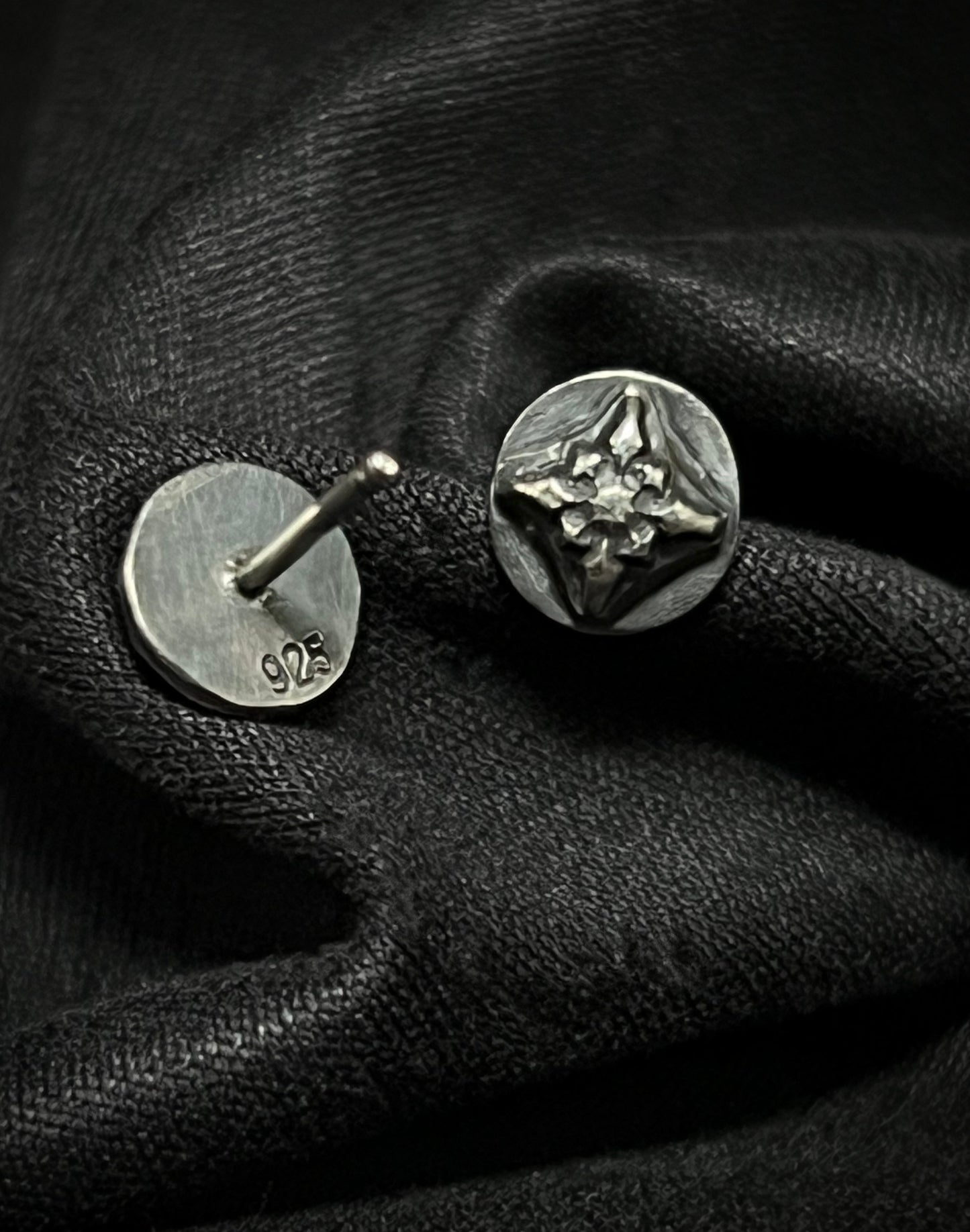 Sterling silver Ember Studs from the Ashborn Collection. The image shows one earring face-up, revealing a polished, raised four-pointed starburst design against a dark patina, and one earring face-down showing the '925' hallmark, both resting on draped black fabric.