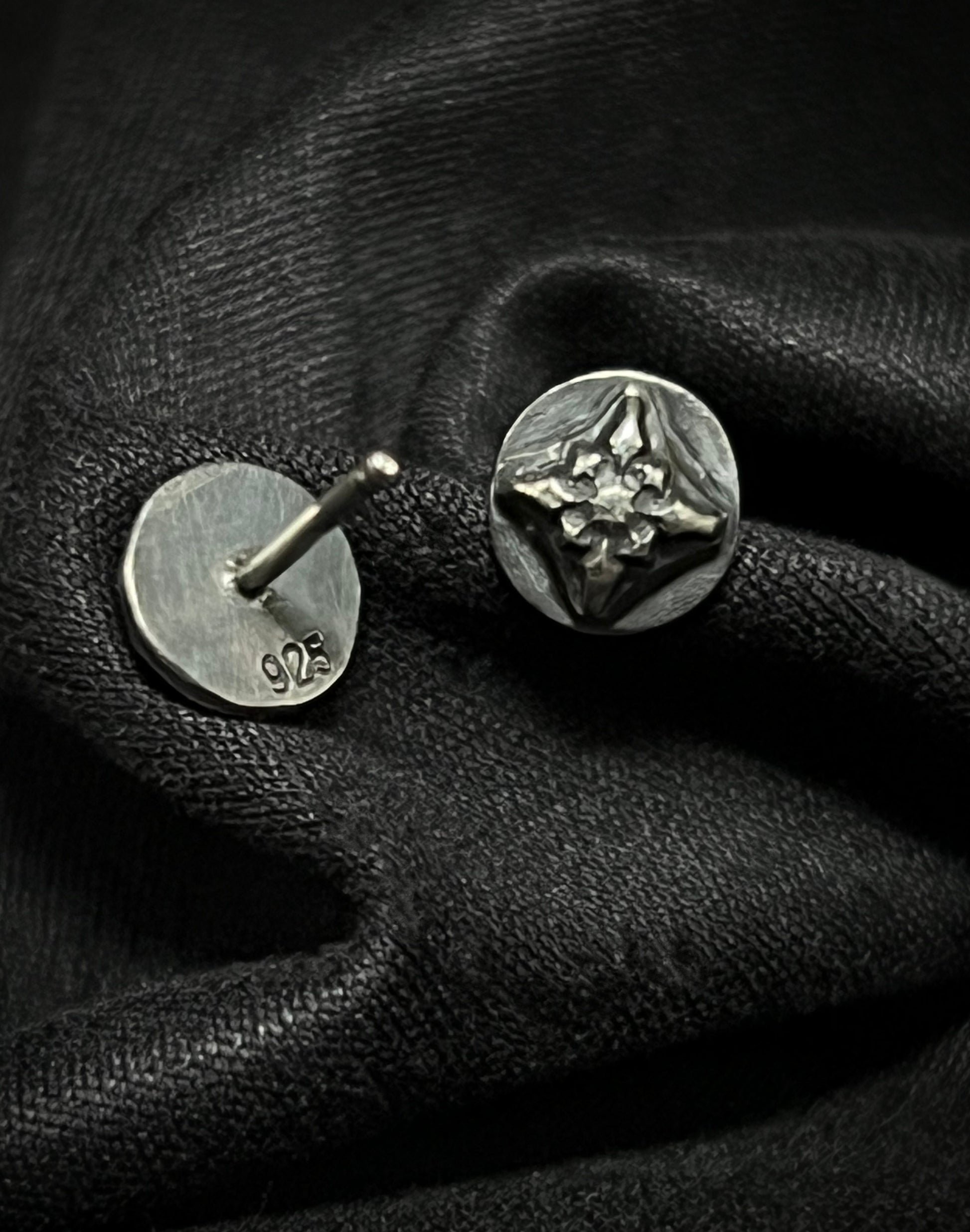 Sterling silver Ember Studs from the Ashborn Collection. The image shows one earring face-up, revealing a polished, raised four-pointed starburst design against a dark patina, and one earring face-down showing the '925' hallmark, both resting on draped black fabric.