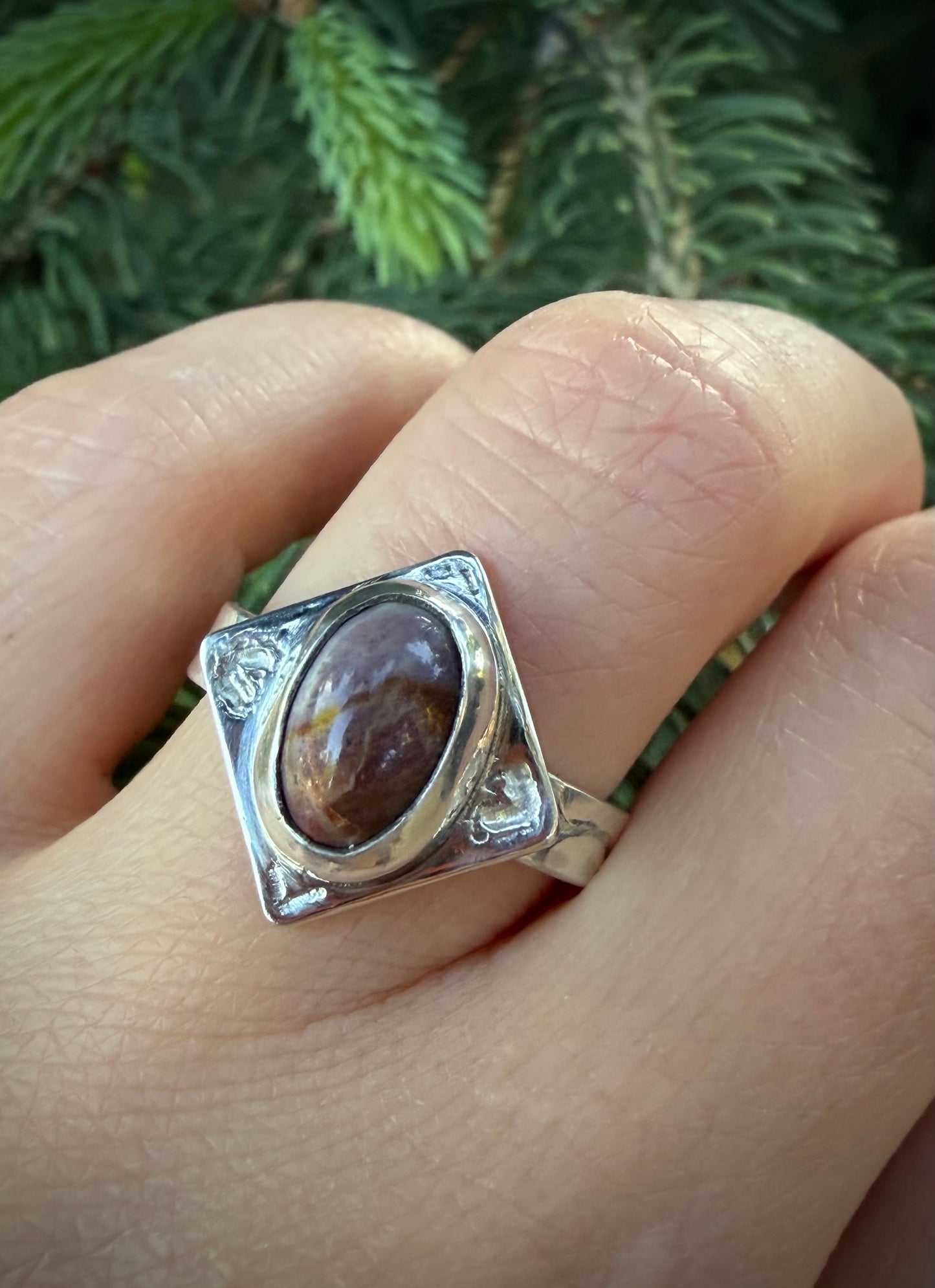 A handcrafted sterling silver Tephra Ring featuring a diamond-shaped plate with engraved heart motifs and a raw, molten texture. The ring is set with a polished, earth-toned agate and worn on a finger against a blurred natural green background.