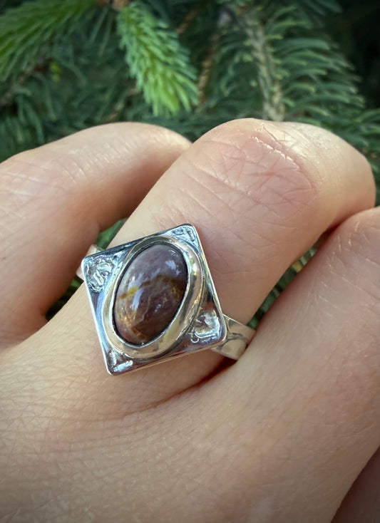 A handcrafted sterling silver Tephra Ring featuring a diamond-shaped plate with engraved heart motifs and a raw, molten texture. The ring is set with a polished, earth-toned agate and worn on a finger against a blurred natural green background.